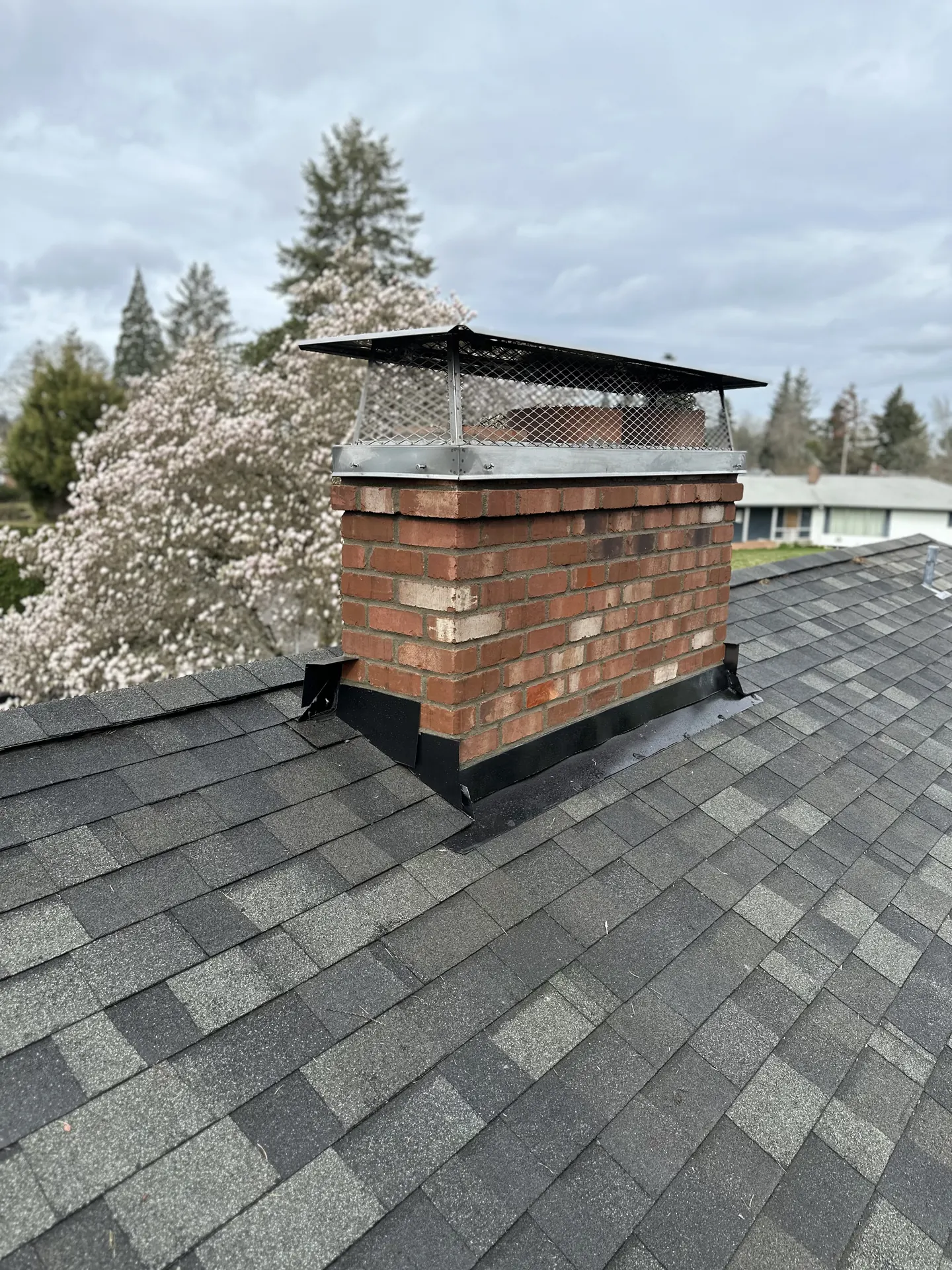 A wide custom black chimney cap on a rebuilt brick chimney against spring blossoms near Gresham, Oregon